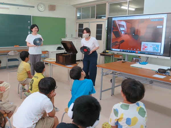 レコード盤を持って蓄音機の解説をする学芸員と話を聞く子ども